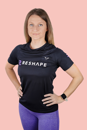 Alex, a personal trainer for women in Dubai, wearing a black Reshape t-shirt and purple leggings, standing confidently against a pink background